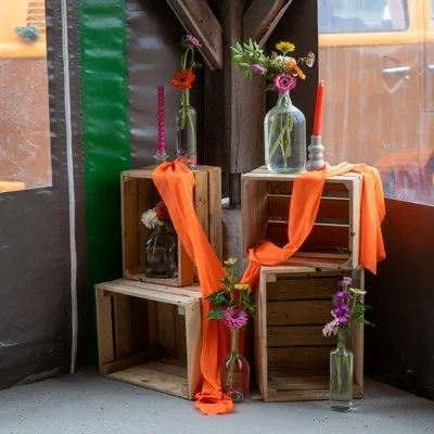 Cagettes en bois drapées de voile orange, avec des fleurs en bouteilles de verre et des bougies