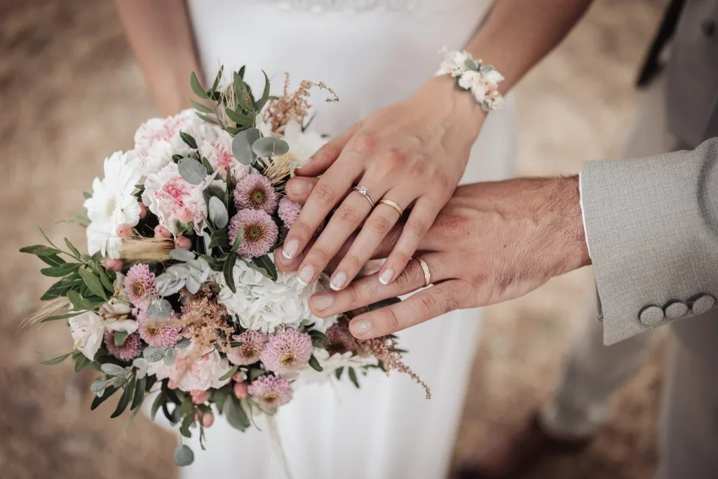 Mains des mariés avec leurs alliances et le bouquet de mariée rose et blanc
